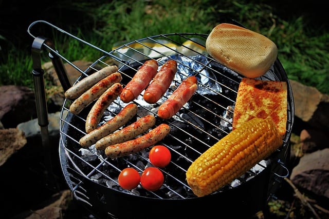 Hiiligrilli täynnä erilaisia makkaroita, maissia, tomaattia ja leipää, grillaus käynnissä puutarhassa.