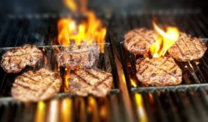 Grillissä kypsyviä hampurilaispihvejä liekkien loimutessa, symboloiden hiili- ja kaasugrillin eroja.