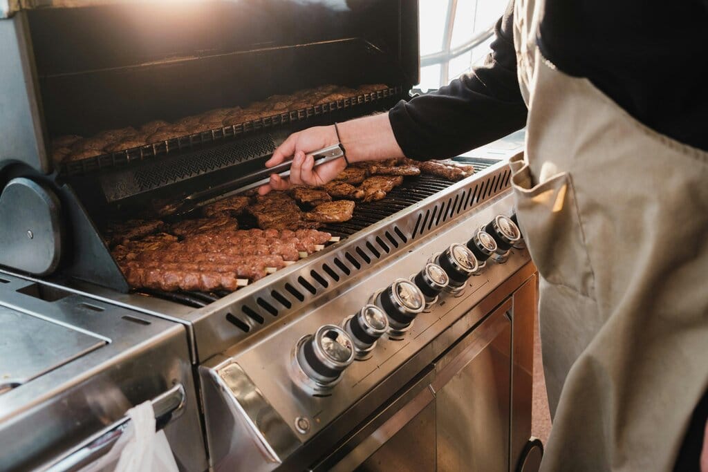 "Henkilö grillaamassa lihaa kaasugrillissä, lähikuva grillin säätimistä ja ritilällä kypsyvistä vartaisista.