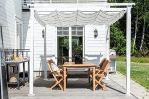 Valkoinen pergola kattaa puisen ruokailuryhmän terassilla, aurinkoinen kesäpäivä ja siististi asennettu varjokangas