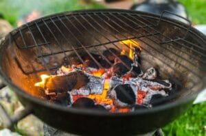 Lähikuva hehkuvista hiilistä ja liekeistä pyöreässä hiiligrillissä ulkona. Kuuma grilli on valmis ruoanlaittoon.
