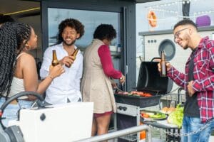 Iloinen ystäväporukka grillaa sähkögrillillä parvekkeella, nauttien ruoasta ja seurasta.