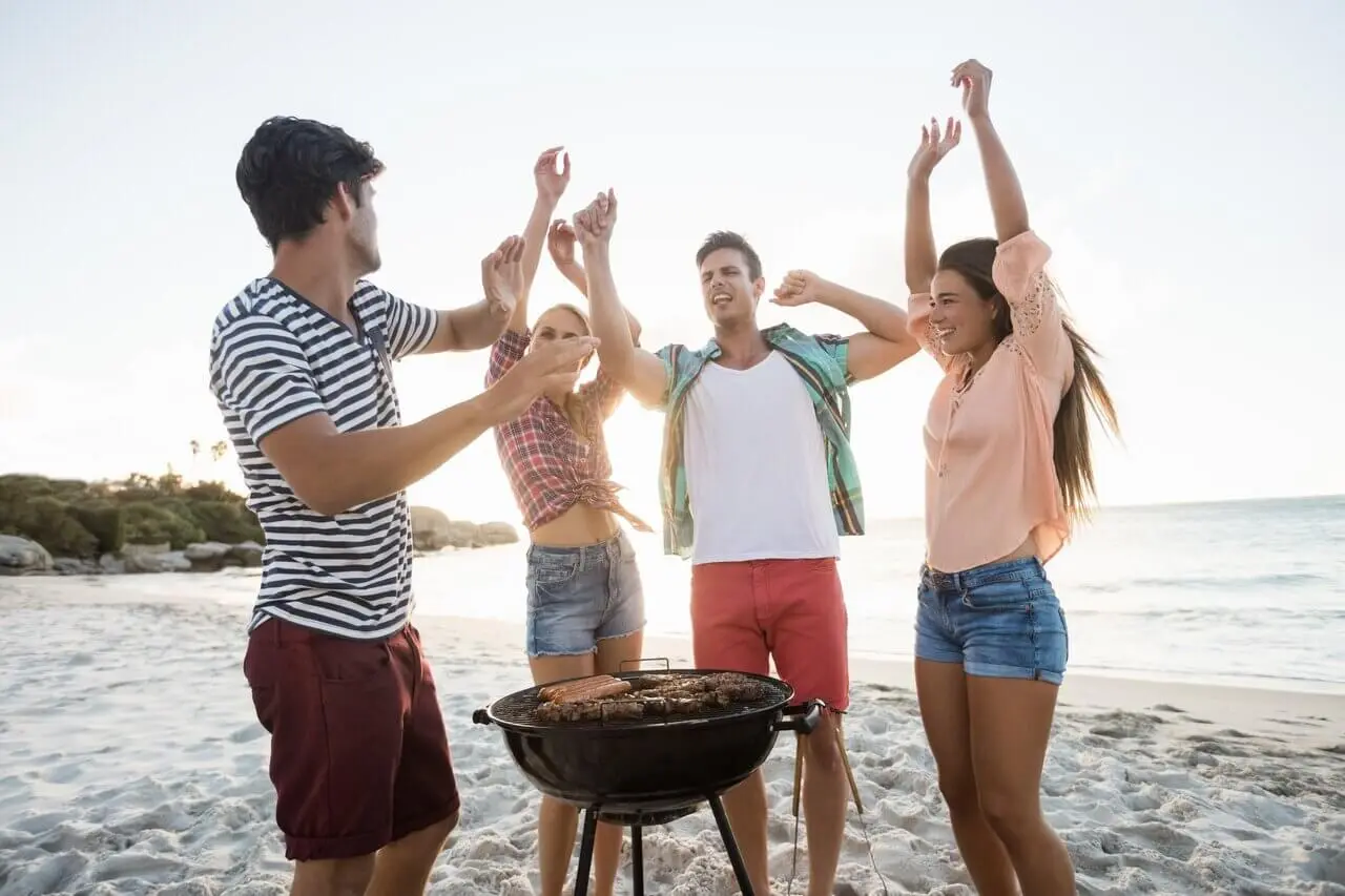 Ystäväporukka rannalla grillin ympärillä matkagrilli käytössä.