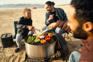 Kolme ihmistä grillaamassa vihanneksia rannalla kannettavalla grillillä – täydellinen esimerkki retkigrillin käytöstä luonnossa.