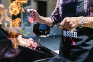 Mies sivelee grillikastiketta lihalle liekkien leimutessa grillissä – esimerkki pienellä kaasugrillillä grillaamisesta vuonna 2025.