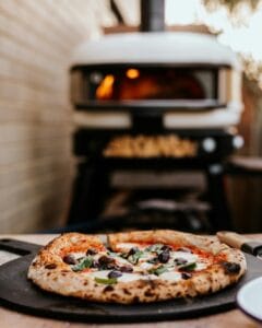 Lähikuva valmiista pizzasta pizzakivellä, taustalla tulikuuma pizzauuni ulkotilassa – täydellinen kotipizzakokemus.