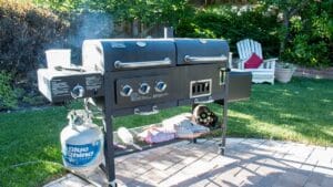 Kaksikammioinen savustin/grilli pihalla, kaasupullo ja tarvikkeita näkyvissä – täydellinen kesäkeittiö savustamiseen ja grillaamiseen.