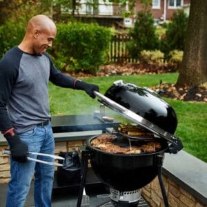 Mies grillaamassa ulkona Weberin mustalla hiiligrillillä, jossa kannen alla useita grillattavia ruokia – liittyy vuoden 2025 Top 3 -hiiligrillivertailuun.