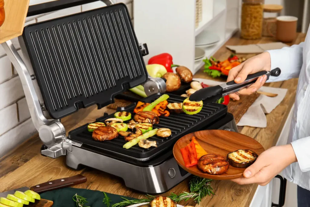 Pöytägrilli keittiötasolla käytössä, kasviksia grillataan paistolevyllä.