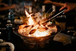 Grillattavaa ruokaa kypsymässä kompaktissa pöytägrillissä liekkien päällä, taustalla illanviettoa – tehokas ratkaisu pieniin tiloihin ja ulkokäyttöön.