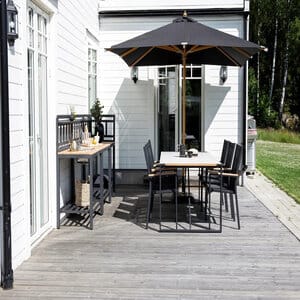 Venture Home Naxos -aurinkovarjo mustalla kankaalla ja luonnonvärisellä rungolla terassin ruokailuryhmän yllä – moderni ja luonnonläheinen ilme.