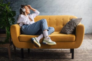 Nuori nainen istuu keltaisella sohvalla kuunnellen musiikkia kuulokkeilla ja hymyillen, sisustuksessa rento ja moderni tyyli.