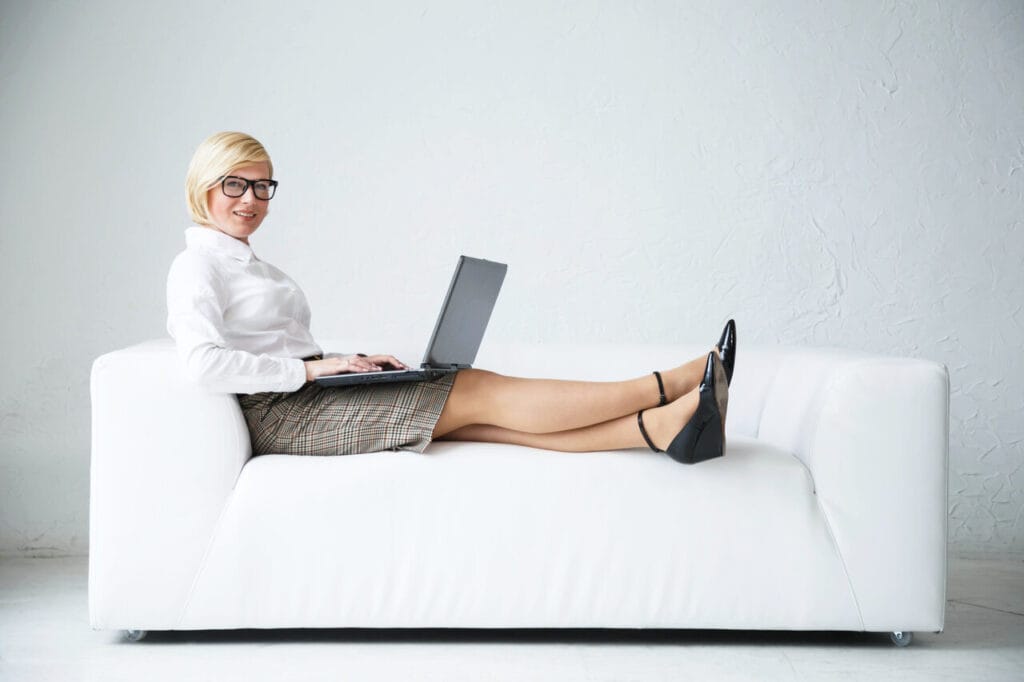Hymyilevä nainen istuu valkoisella nahkasohvalla jalat suorana ja työskentelee kannettavalla tietokoneella, taustalla minimalistinen vaalea sisustus.
