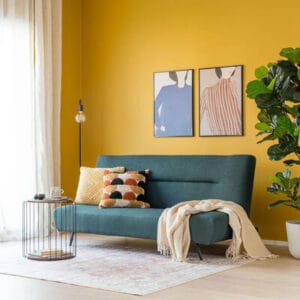 Tummanvihreä Furry 3-istuttava vuodesohva keltaisella seinällä, koristetyynyt ja sivupöytä täydentävät kokonaisuuden