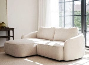 Vaalea Rowico Home Norris mini divaanisohva kaarevalla muotoilulla, sijoitettuna valoiseen olohuoneeseen suurten ikkunoiden eteen – pieni ja moderni divaanisohva