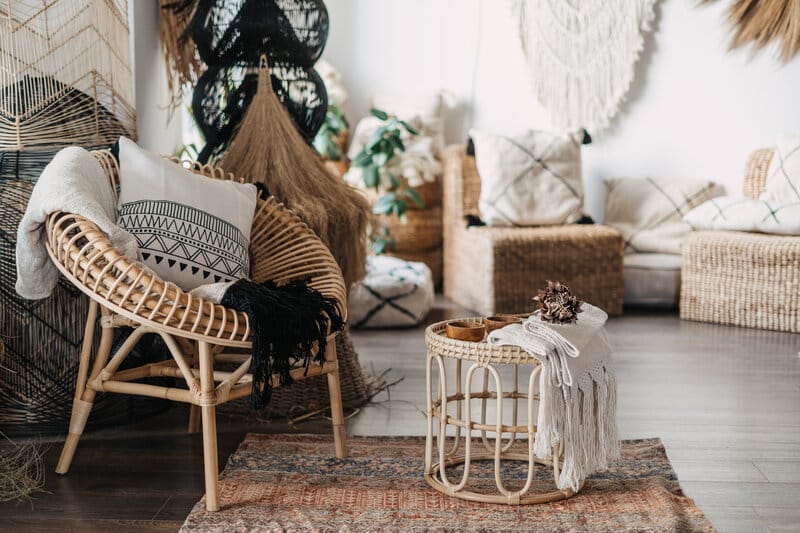 Boho-tyylinen olohuone rottinkisella nojatuolilla, punasävyisellä matolla ja luonnonmateriaaleista valmistetuilla sisustuselementeillä