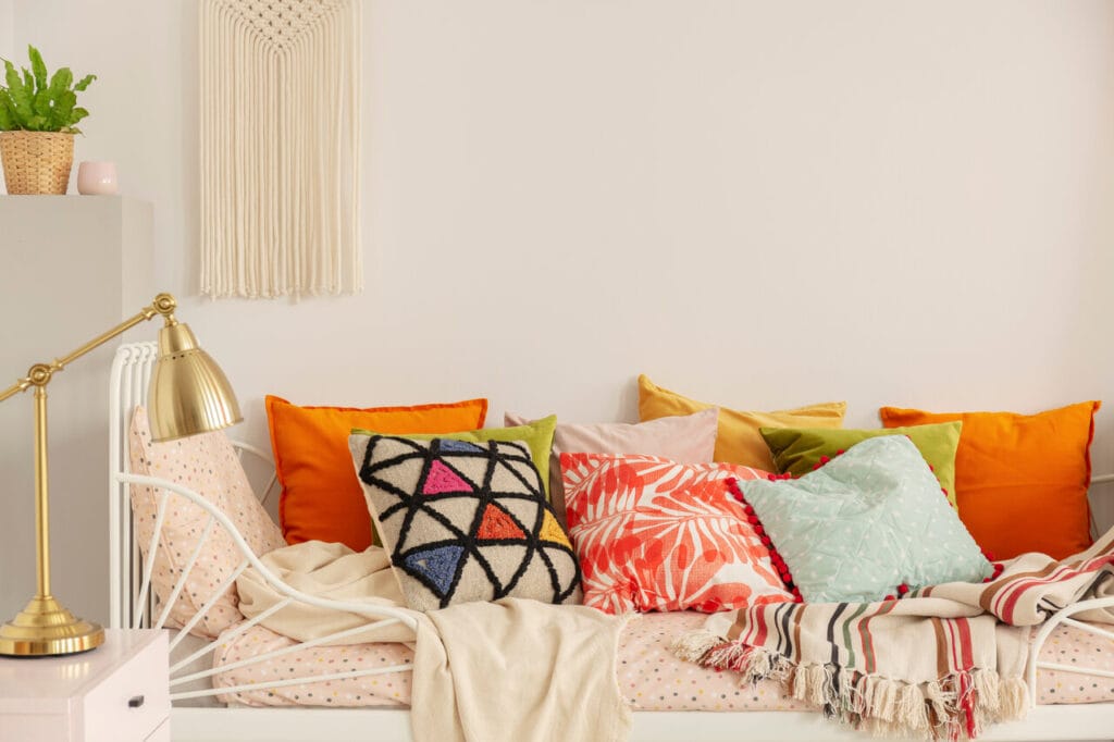 Boho-tyylinen päivävuode täynnä värikkäitä koristetyynyjä ja pehmeitä torkkupeittoja valkoista seinää vasten