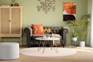 Tumma Chesterfield-tyylinen 2-istuttava sohva vihreäksi maalatussa olohuoneessa, koristeena oranssi tyyny ja kukkia pöydällä