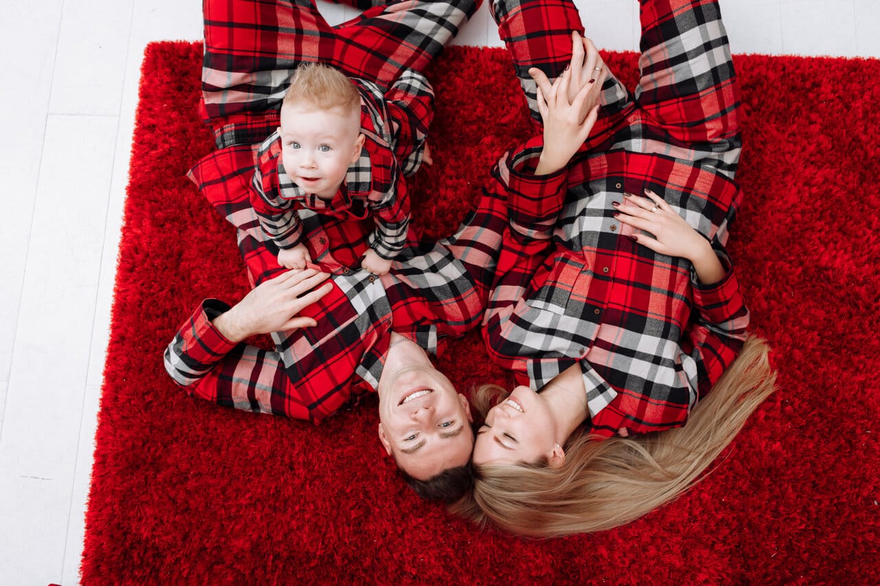 Perhe punaisissa ruutupyjamoissa makaa punaisella pehmeällä matolla ja hymyilee