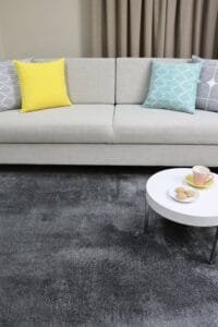 Mustanharmaa Rex-nukkamatto modernissa olohuoneessa, jossa on vaalea sohva ja värikkäitä tyynyjä