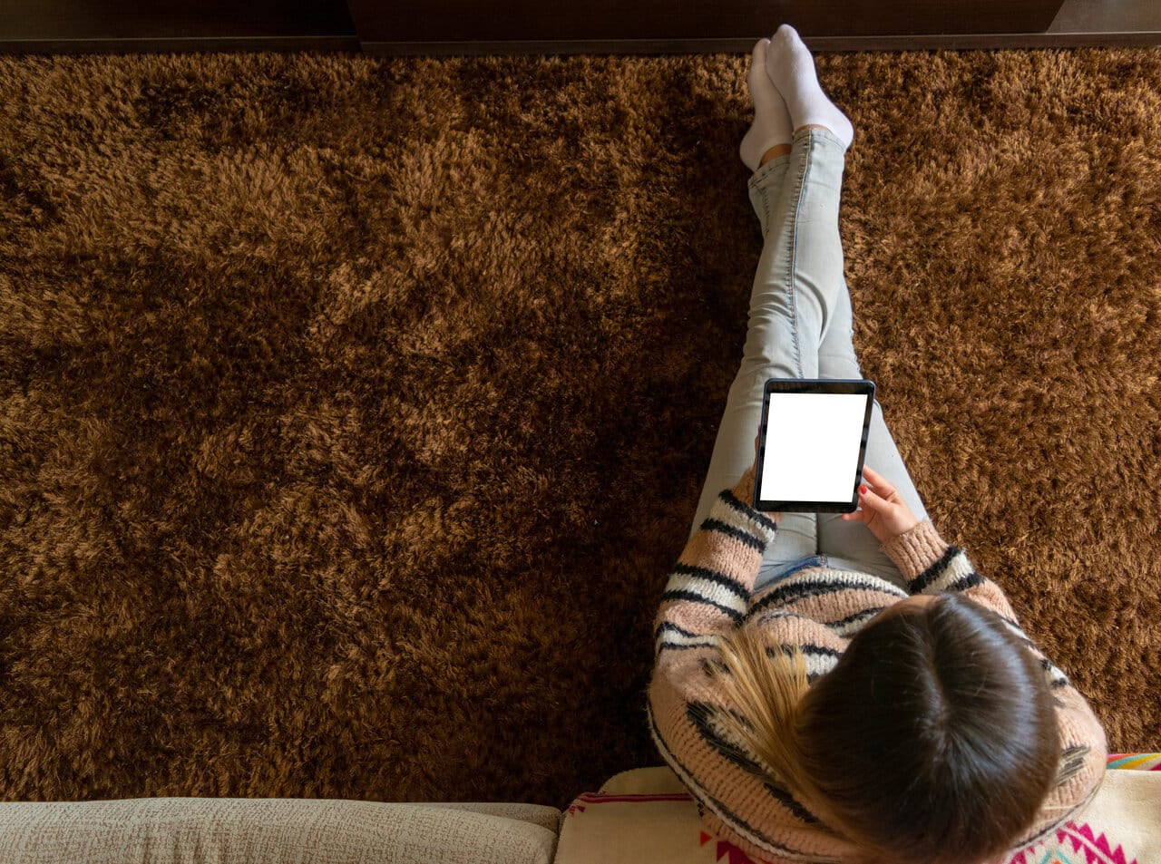 Nainen istuu ruskealla ryijymatolla ja käyttää tablettia olohuoneessa