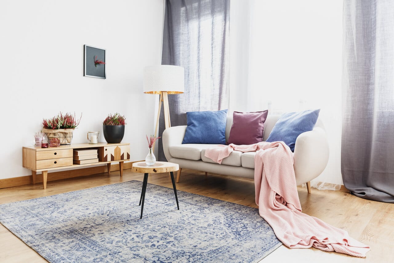 Vaaleansininen kuviollinen matto olohuoneessa, jossa beige sohva, sinisiä ja liiloja tyynyjä sekä vaaleanpunainen viltti