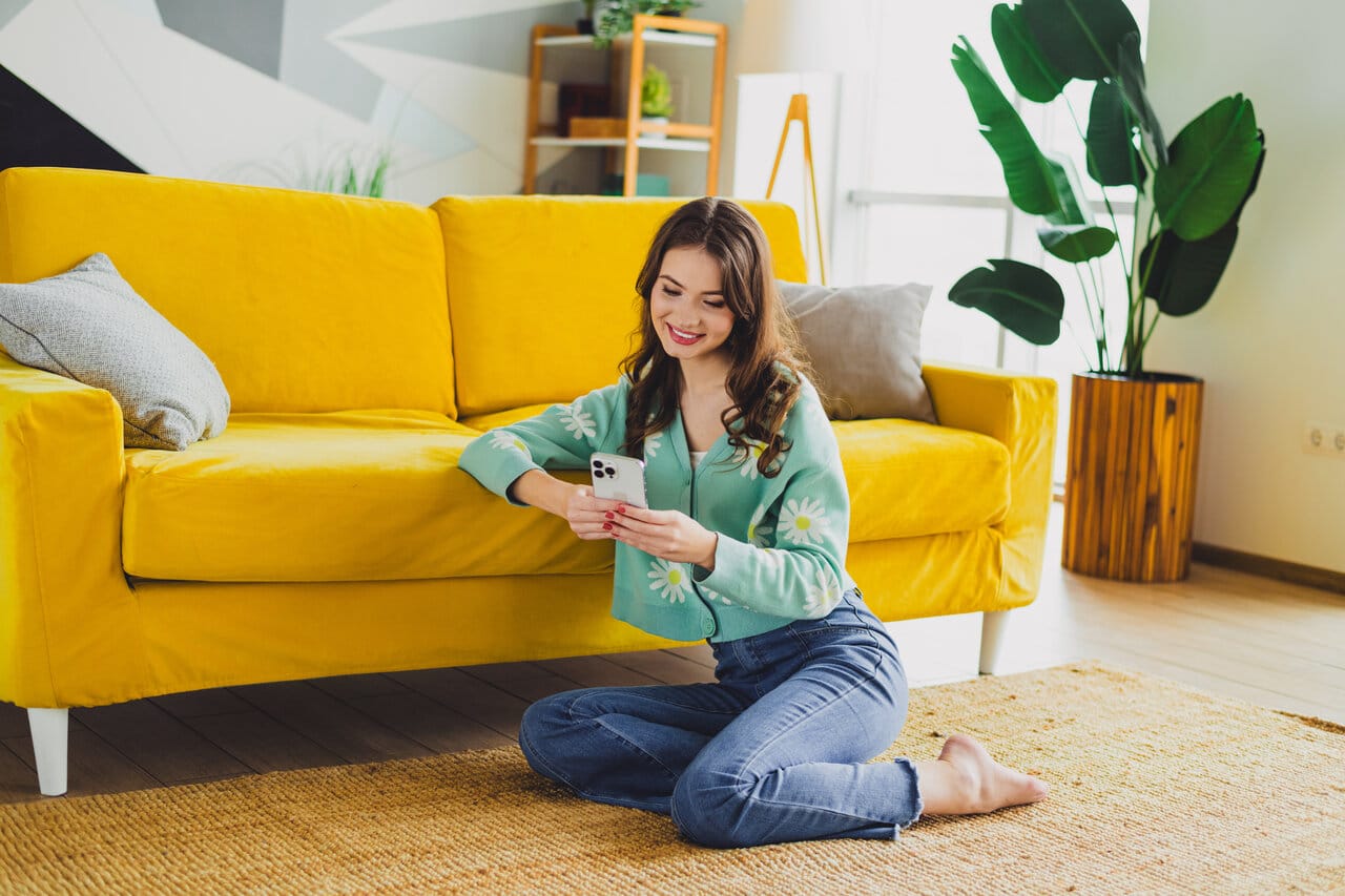 Nainen istuu keltaisen sohvan edessä keltaisen maton päällä, käyttää puhelinta valoisassa olohuoneessa, taustalla vihreitä kasveja