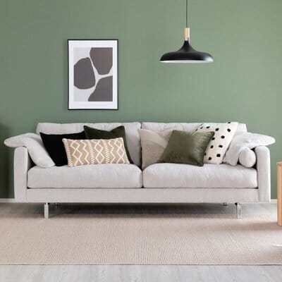 Montesano 3-istuttava beige sohva vihreää seinää vasten, koristeltu tyynyillä ja modernilla valaisimella