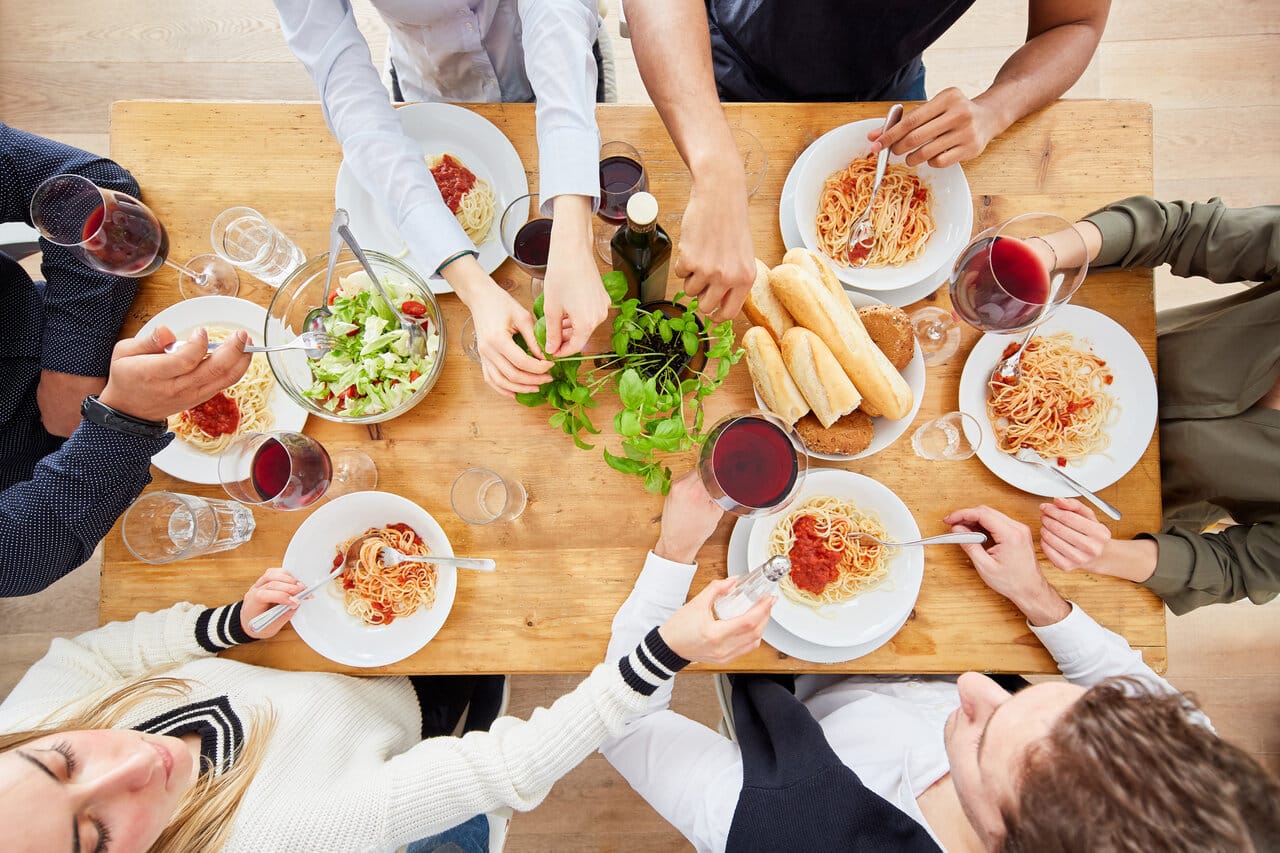 Ilmakuva kuudesta ihmisestä syömässä pastaa ja salaattia puisen ruokapöydän ääressä, viinilasit ja leipäkori pöydällä