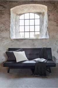 Musta Venture Home Bodil -vuodesohva vanhassa kiviseinäisessä huoneessa, jossa on kaari-ikkuna ja sisustustyynyjä