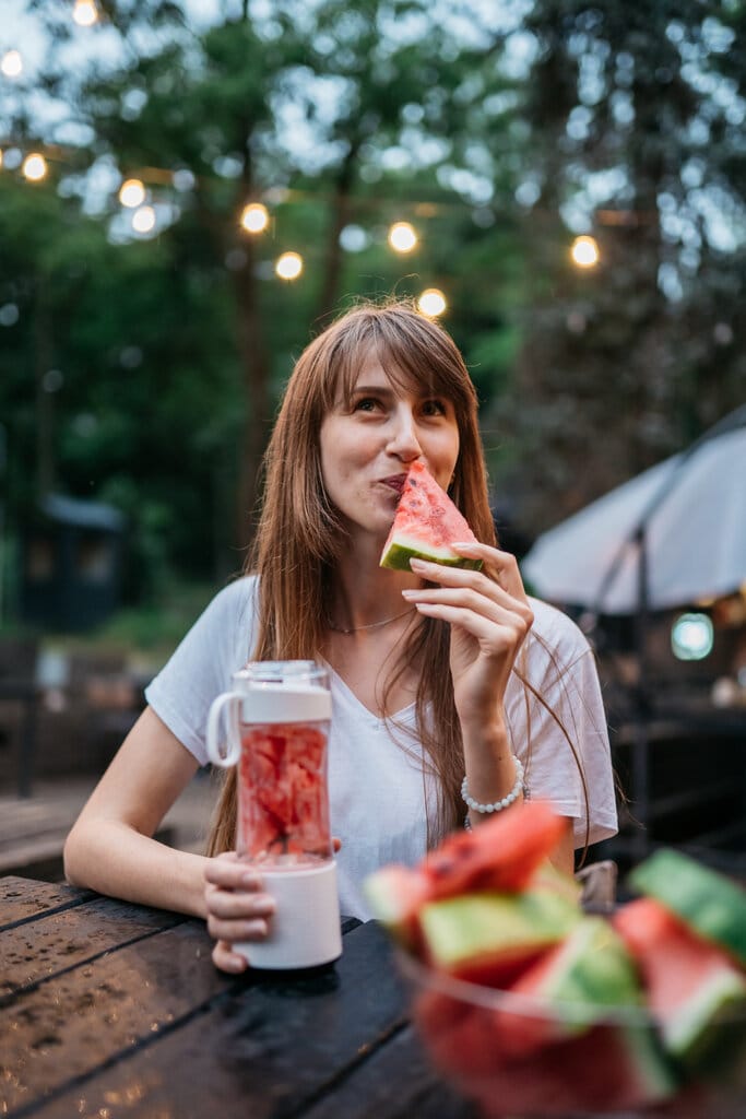 Nainen istuu ulkona ja käyttää kannettavaa tehosekoitinta, pitelee smoothiejuomaa ja vesimeloniviipaletta.