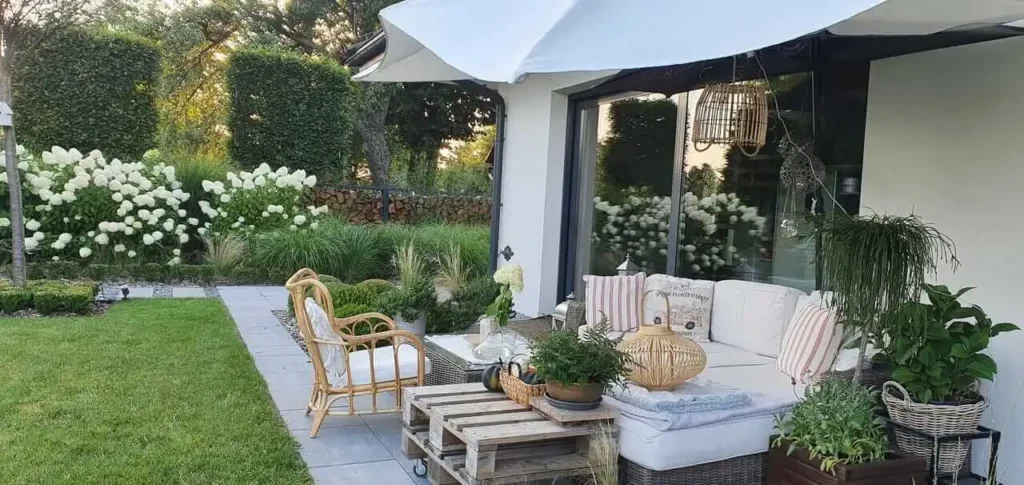 Tyylikäs ja vehreä terassi, jossa on puutarhakalusteita, aurinkovarjo ja istutuksia, moderni koti taustalla.